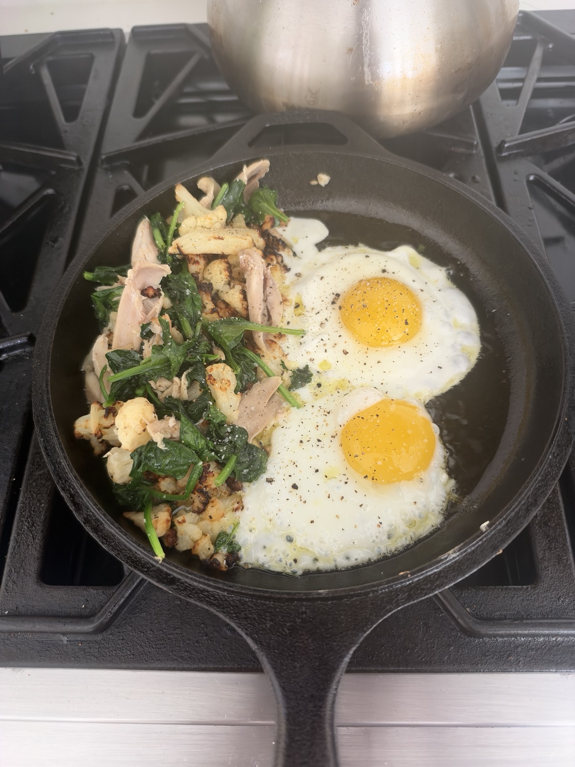 Chicken, roasted cauliflower and spinach reheating beside two eggs in a cast-iron skillet