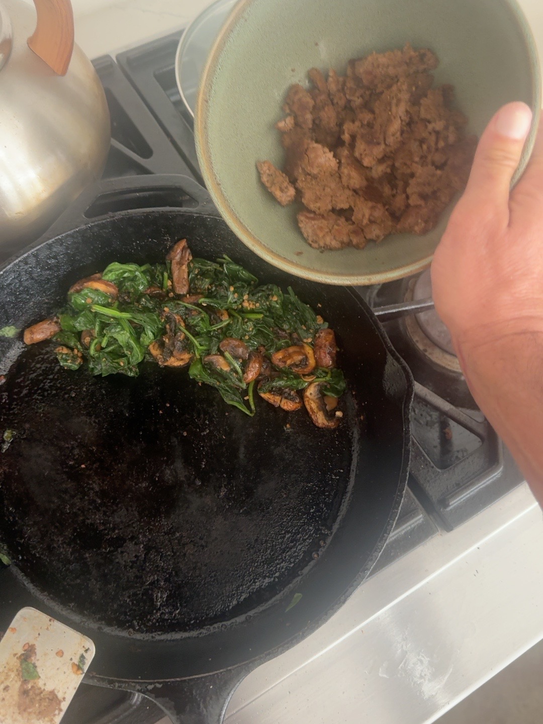 A bowl of cooked ground beef held above the skillet of spinach and mushrooms